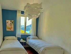 A room with two beds, a window with curtains, a painting on the wall, and a view of mountains. - View over Lake Caldonazzo with large terrace (pergine valsugana)