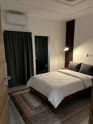 A bedroom with a bed, a bedside lamp, a window with curtains, and an air conditioning unit.