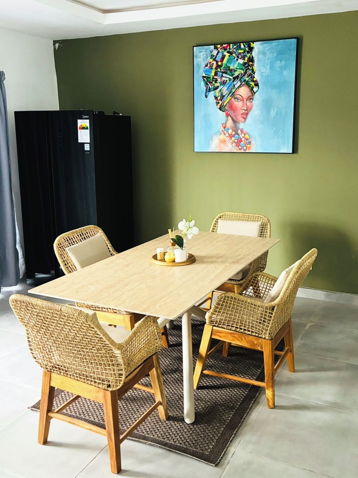 Un coin repas avec une table et quatre chaises en osier, un tableau accroché au mur et un rideau noir en arrière-plan.