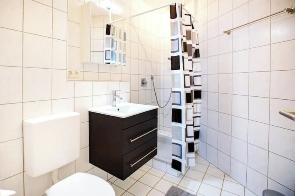 Ein modernes Badezimmer mit Toilette in Weiß, einem schwarzen Waschtisch mit Spiegel und einer Dusche mit weiß-schwarzen Fliesen.
