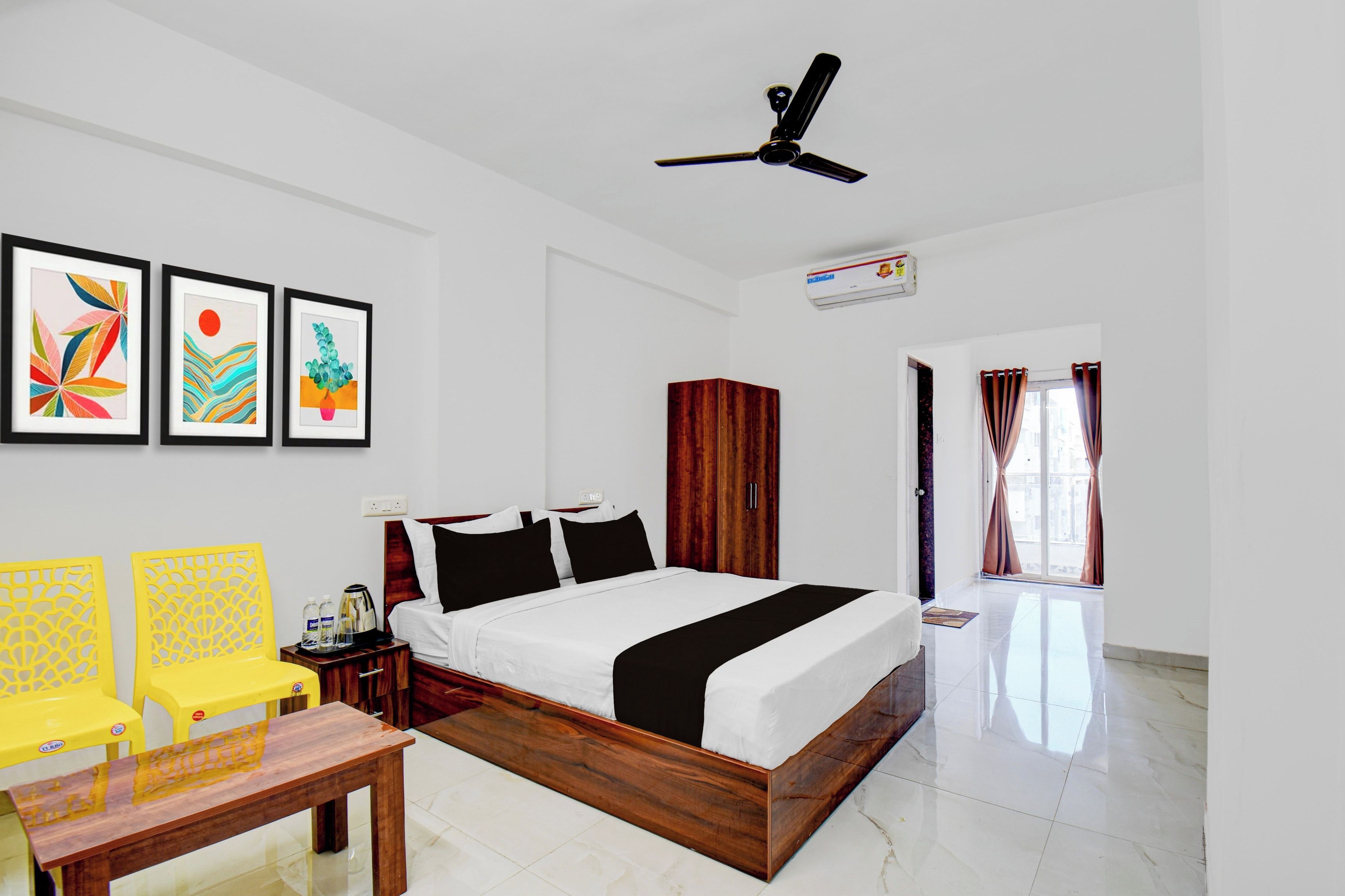 A hotel room with a bed, a desk, chairs, artwork on the wall, and a ceiling fan.
