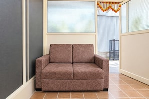 A brown sofa in a corner of a room with tiled flooring and a window with blinds.