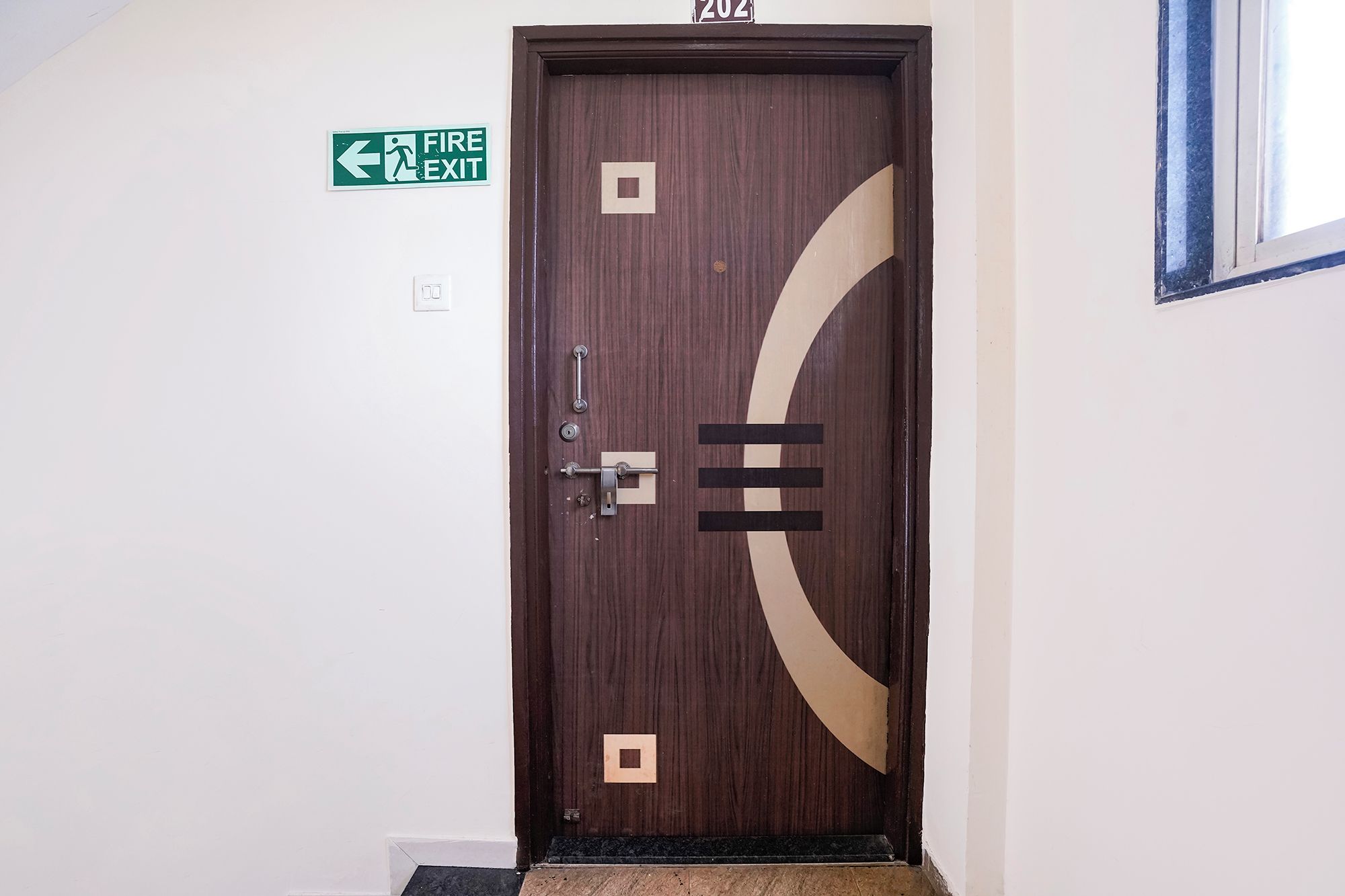 Una puerta con un cartel de salida de emergencia encima, el número '202' en la pared y una ventana a la derecha.