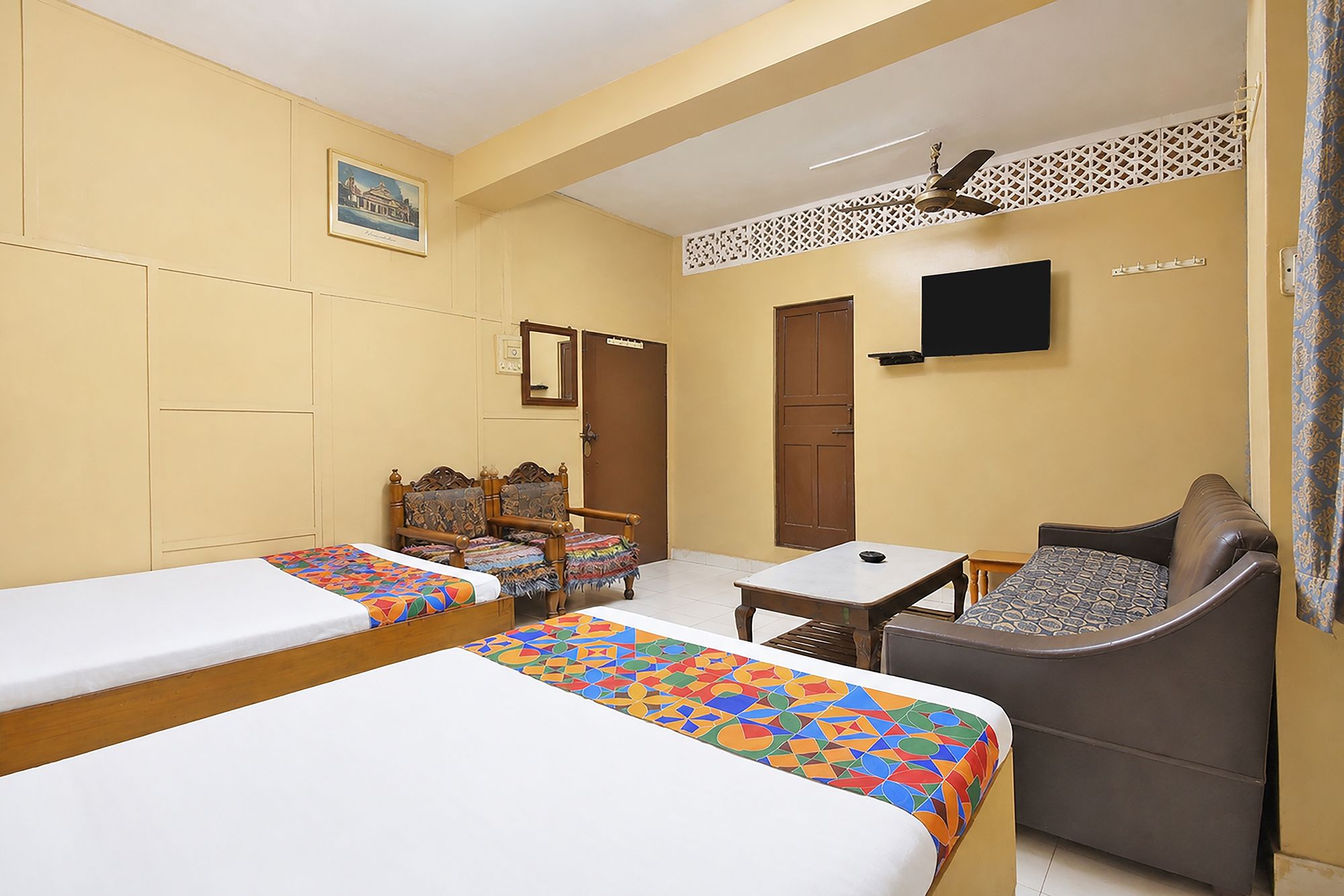 A hotel room with two single beds, a sofa, a small table, a television, and a ceiling fan.