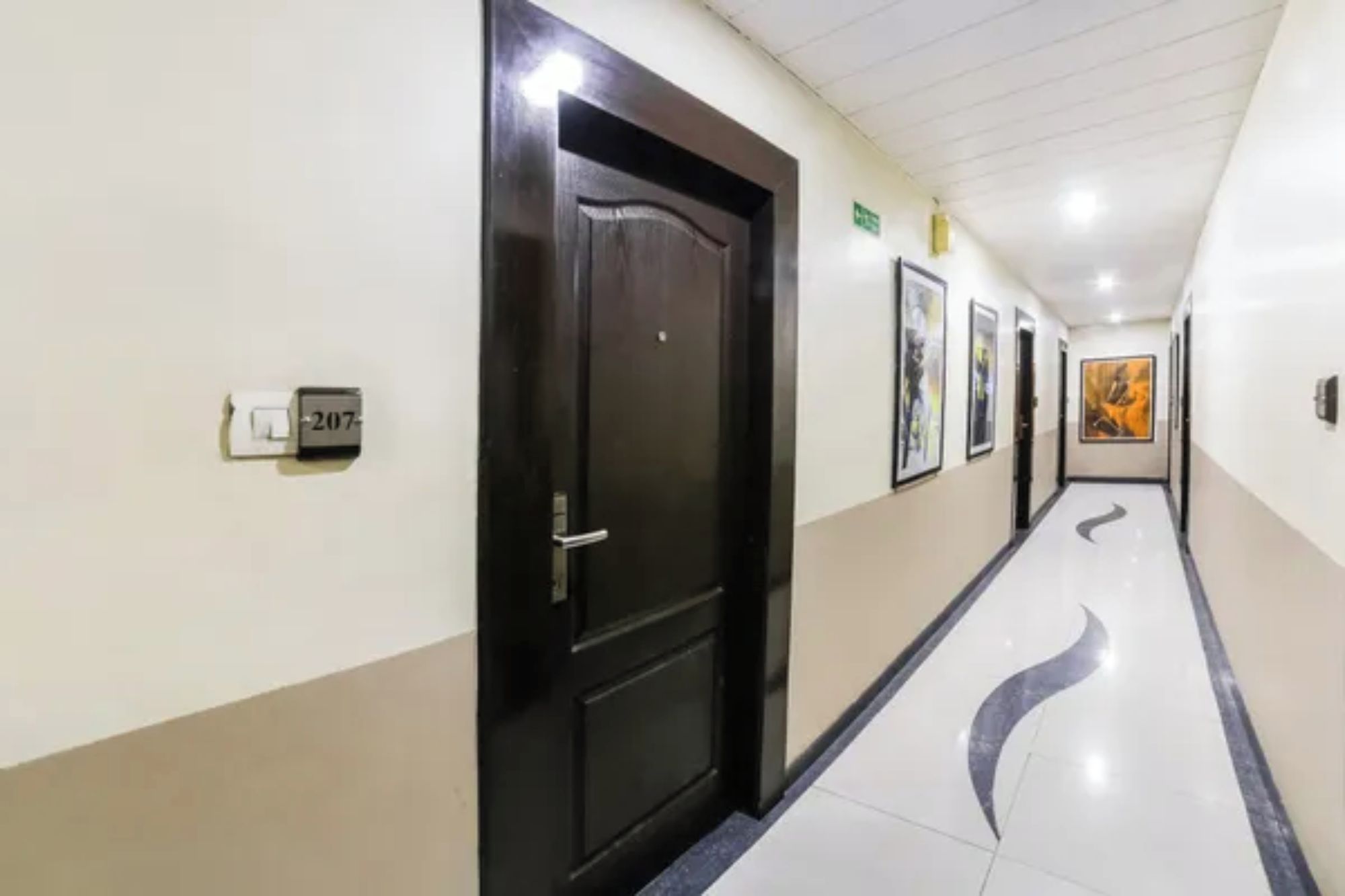 Un pasillo con una puerta negra, un cartel de número de habitación y cuadros enmarcados en la pared.