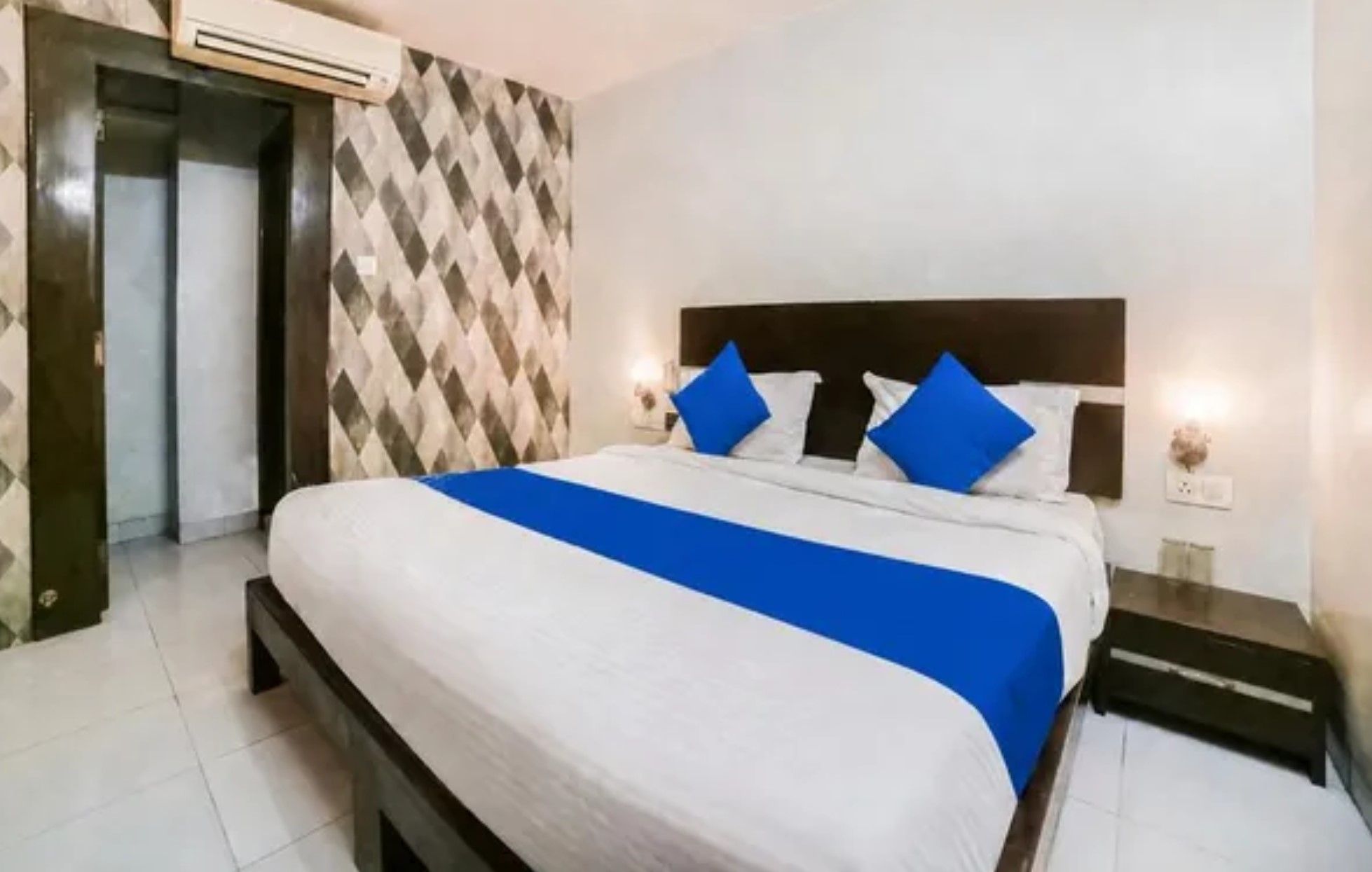 A hotel room with a bed, a nightstand, a wall-mounted air conditioner, and a patterned wall.