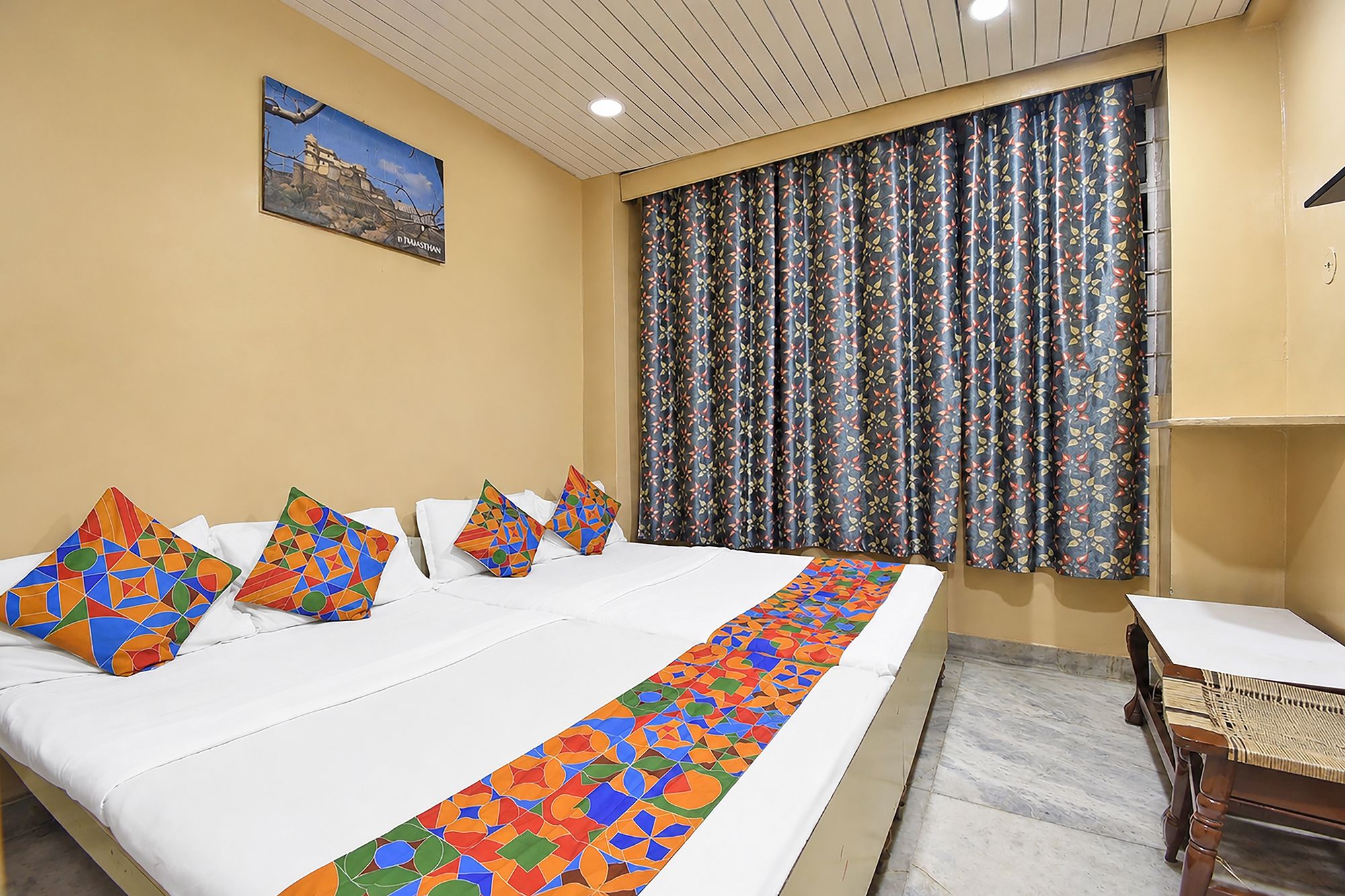 A hotel room with a bed, pillows, a table, a chair, a window with patterned curtains, and a painting on the wall.