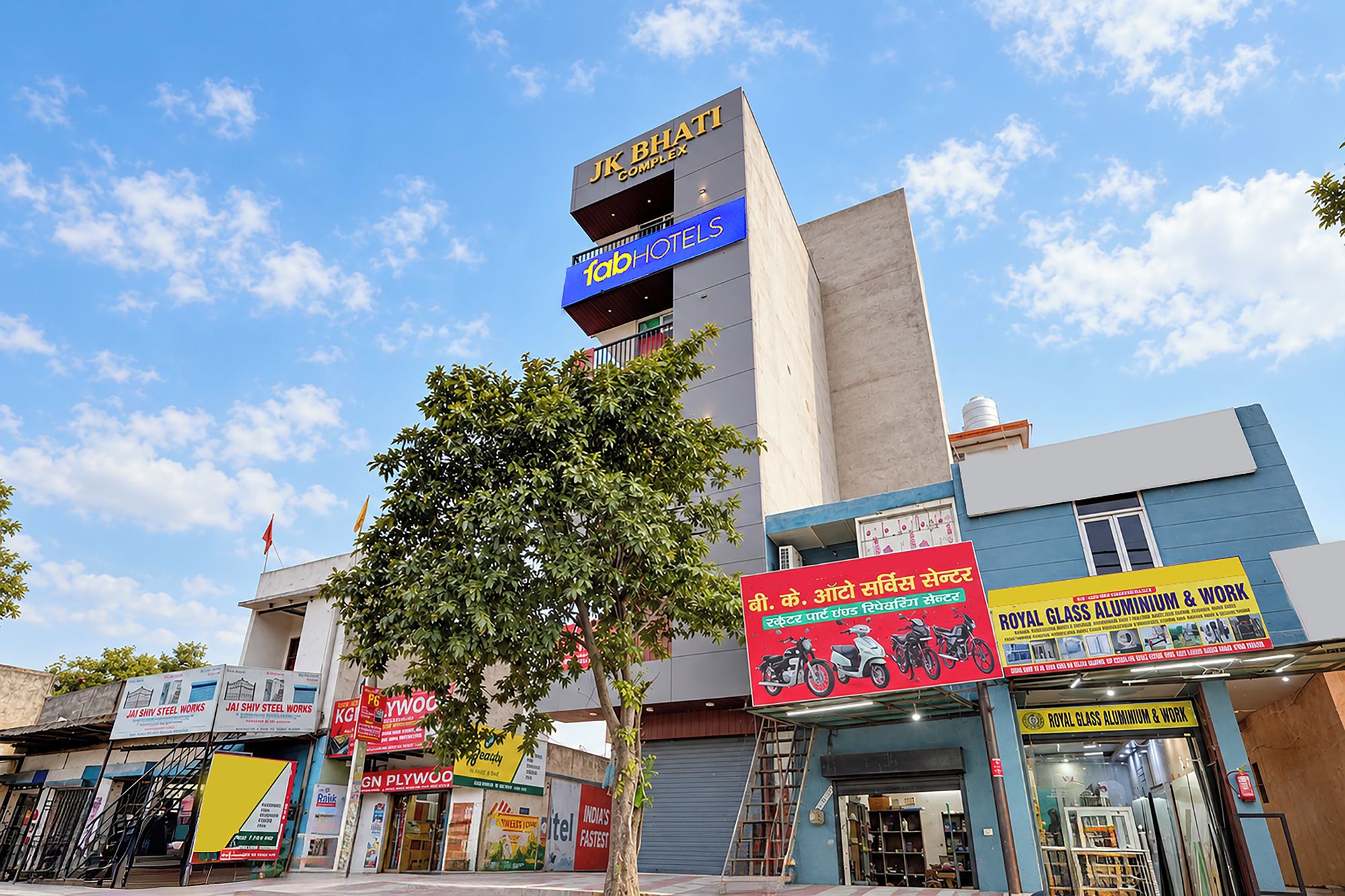 Um edifício comercial com várias fachadas, incluindo um hotel chamado 'Jk Bhati Complex' e uma placa para 'Royal Glass Aluminium & Work'.