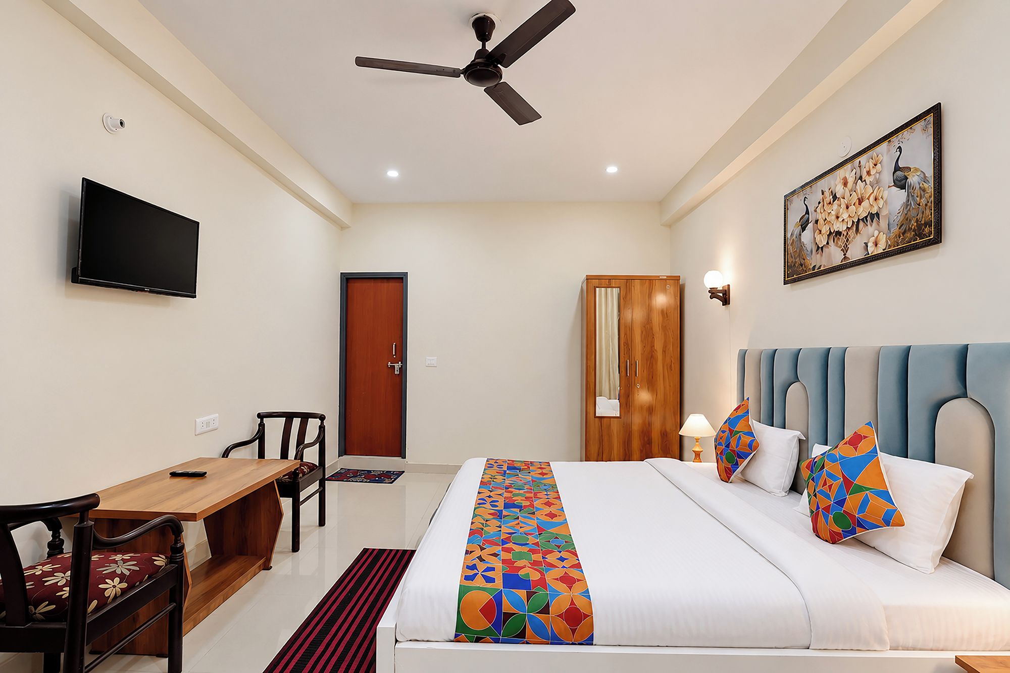 Quarto de hotel com cama, escrivaninha, cadeira, televisão, ventoinha no teto e um quadro na parede.