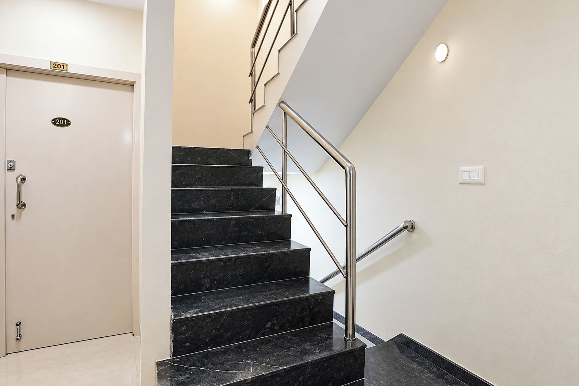 Uma escadaria de mármore preto com corrimãos de metal, uma porta branca com placa de número e um interruptor de luz na parede.