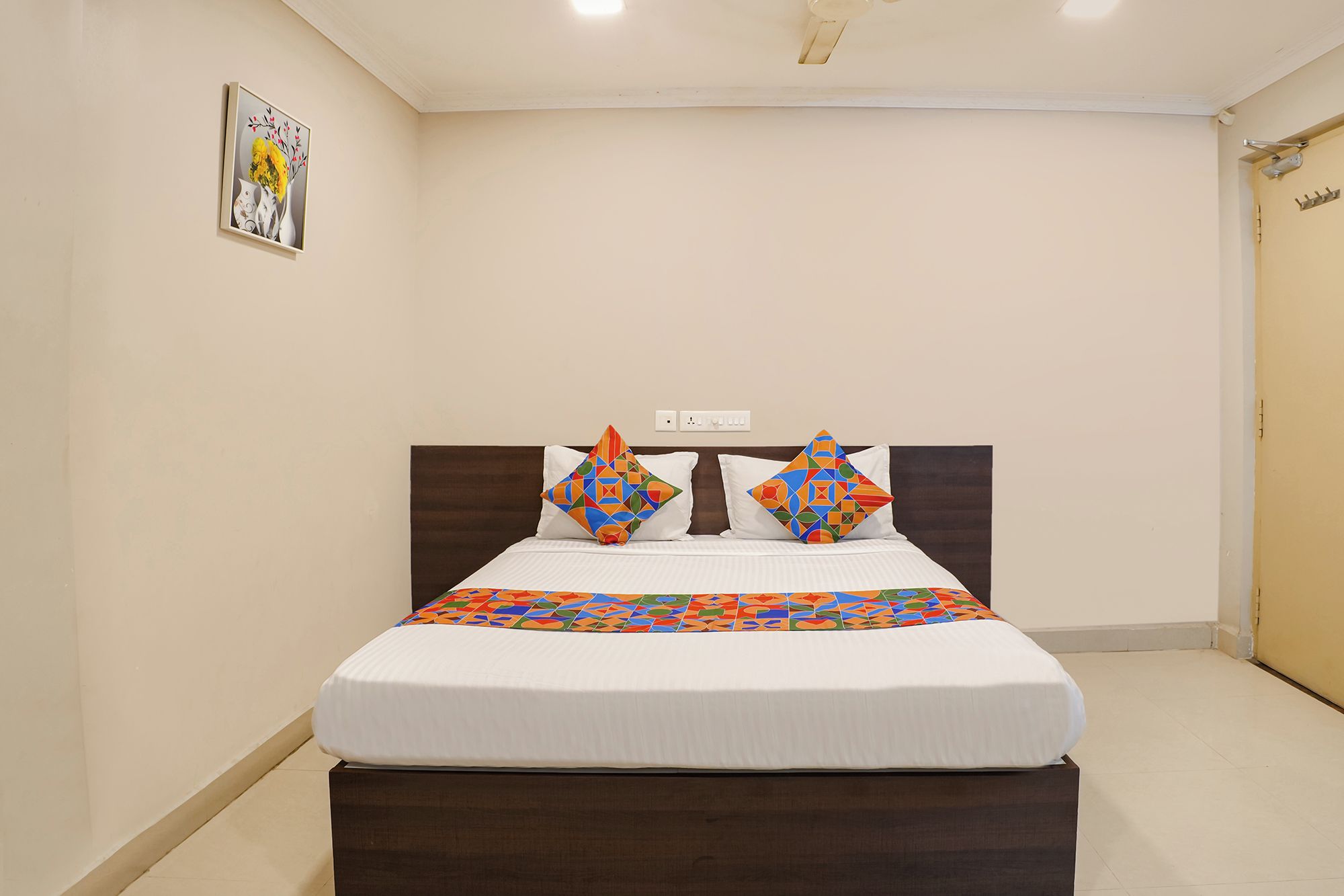 A bed with a patterned bedspread, two pillows with geometric designs, a wooden headboard, and a wall-mounted light fixture.