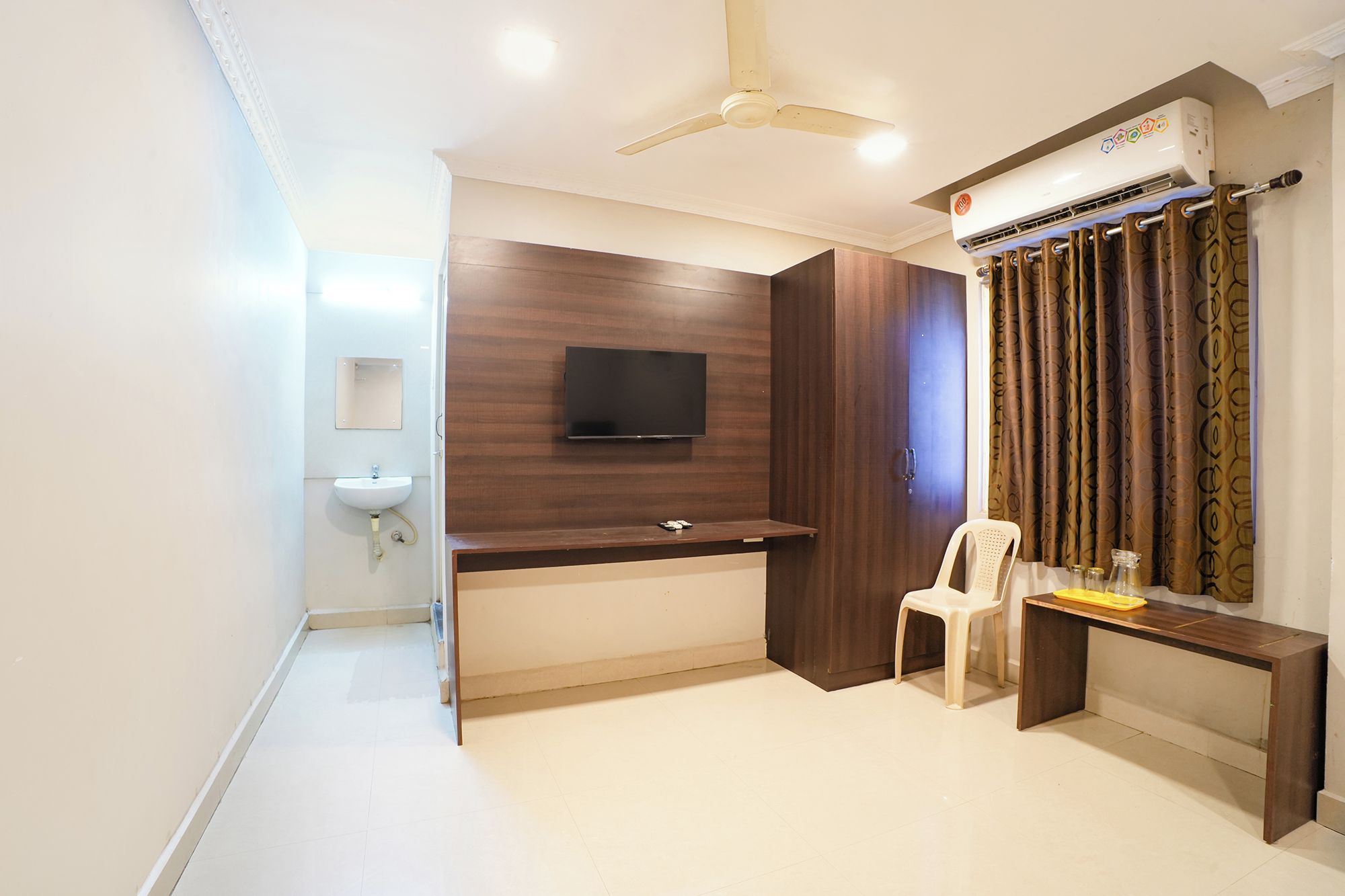 A room with a flat-screen TV mounted on a wooden wall, a ceiling fan, an air conditioning unit, a white sink, and a small table with a chair.