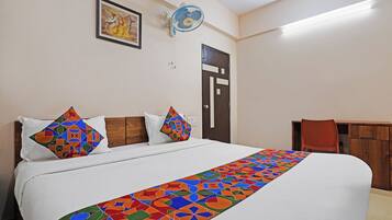 A hotel room with a bed, pillows, a colorful bedspread, a wall-mounted fan, a door, and a small table with a chair.