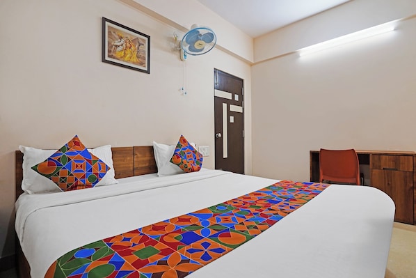 Una habitación de hotel con una cama, almohadas, una colcha colorida, un ventilador de pared, una puerta y una mesa pequeña con una silla.
