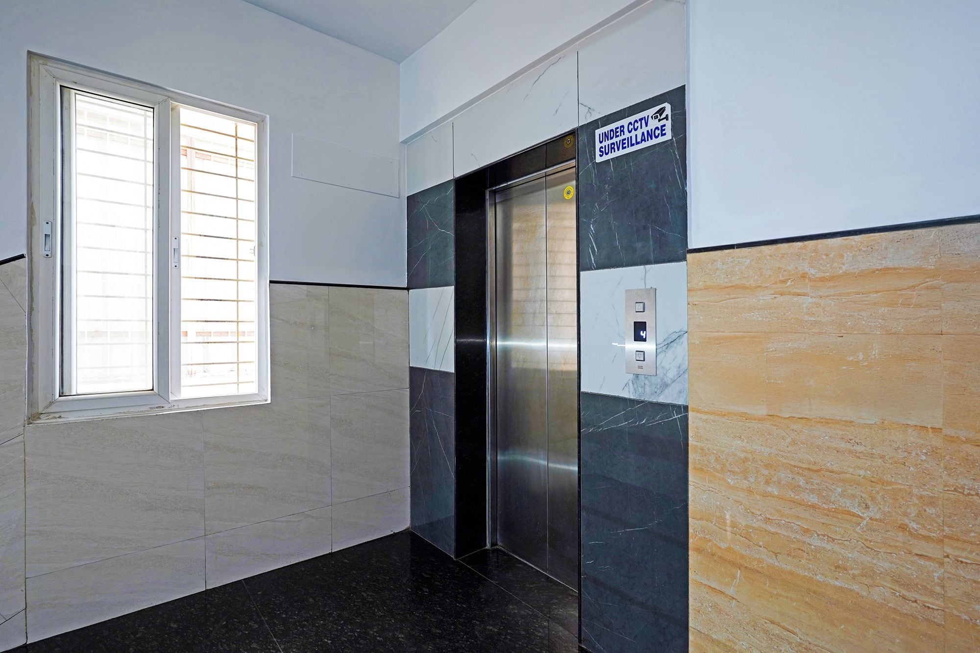 An elevator with a sign indicating CCTV surveillance, a window with blinds, and a marble floor.
