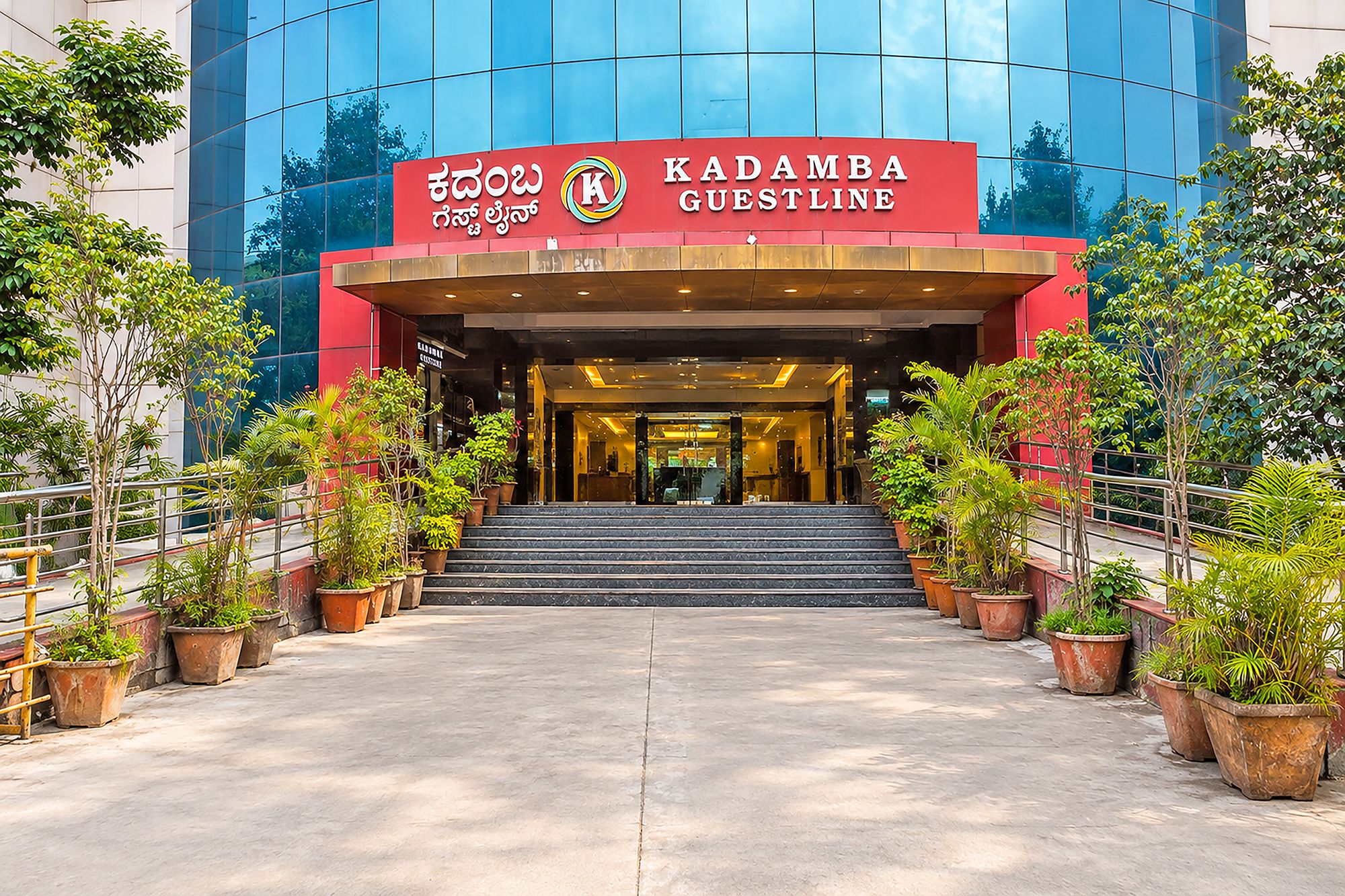 La entrada al hotel Kadamba Guestline, con una fachada moderna de vidrio, un dosel de acceso rojo y una escalera que conduce a la recepción.