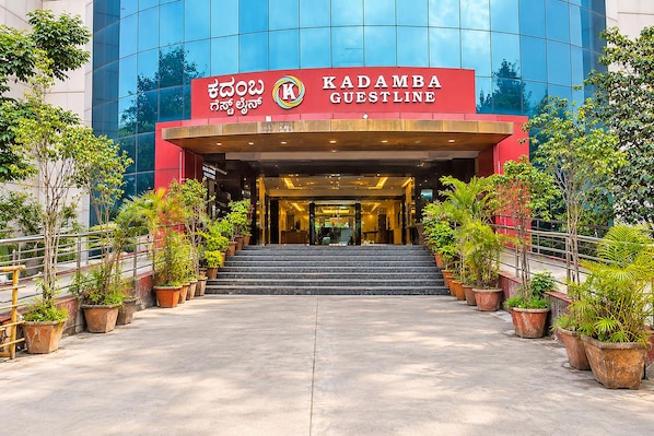 La entrada al hotel Kadamba Guestline, con una fachada moderna de vidrio, un dosel de acceso rojo y una escalera que conduce a la recepción.