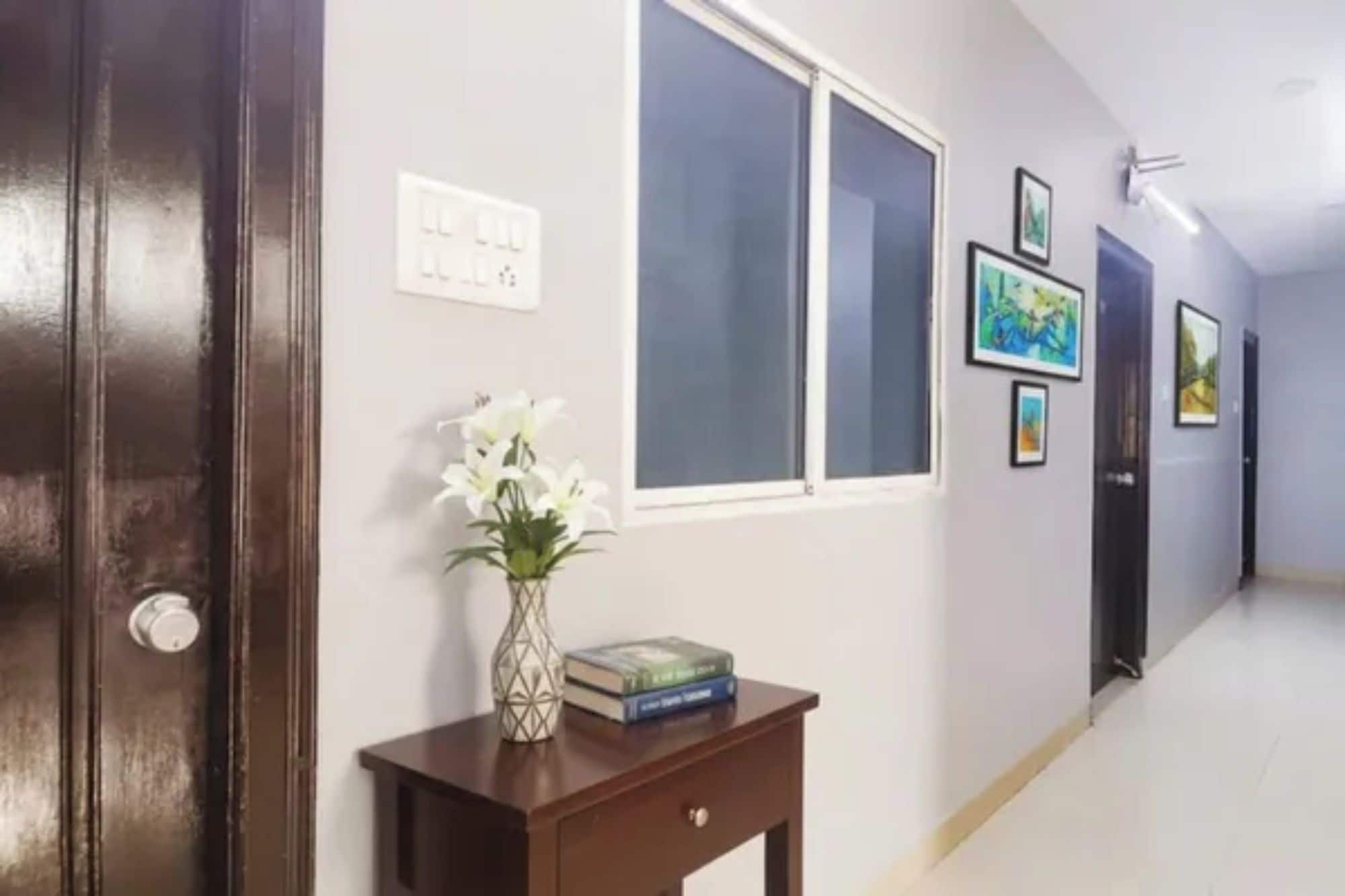 Un couloir avec une porte en bois, une fenêtre, une petite table sur laquelle se trouvent un vase et des livres, et des œuvres d’art encadrées accrochées au mur.