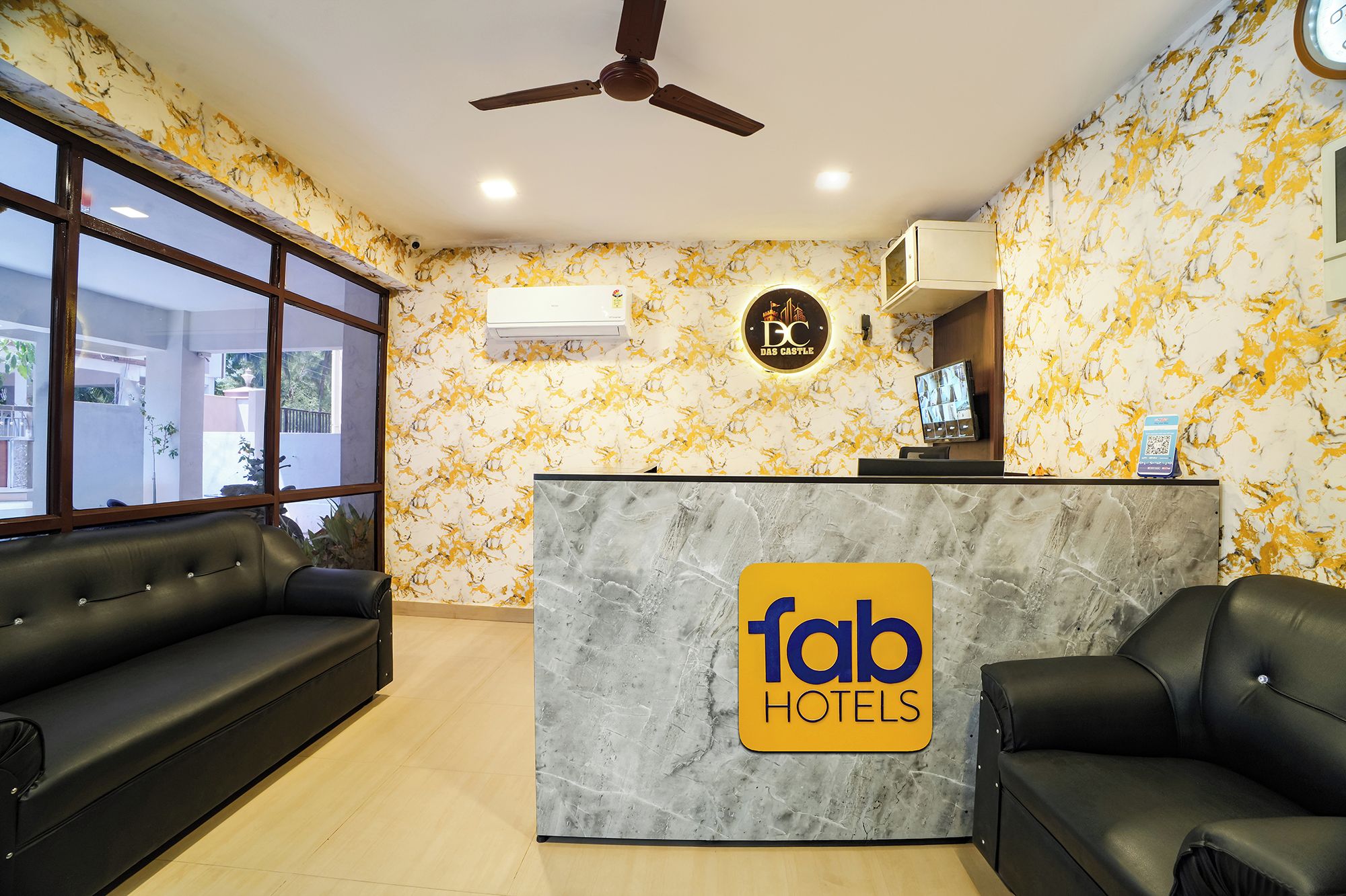 A hotel reception area with a marble counter, two black sofas, a ceiling fan, and a large window.