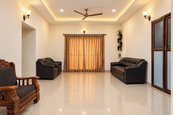 A spacious living room with two leather sofas, a wooden armchair, a ceiling fan, and a large window with curtains.