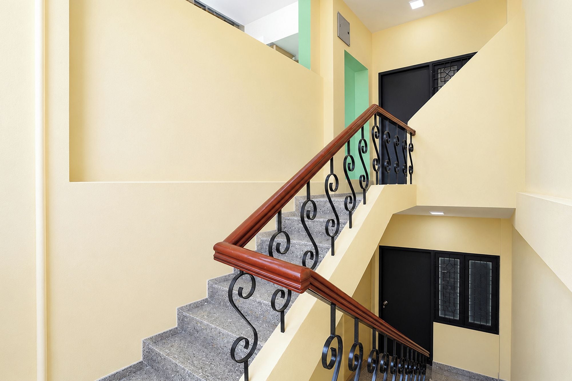 Una escalera con barandilla de madera y barandales de metal negro, que conduce a un rellano con una puerta negra y una ventana con celosía decorativa.
