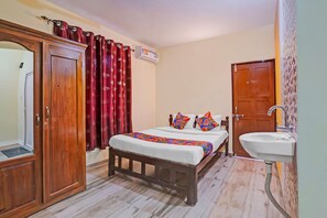 A bedroom with a wooden wardrobe, a bed with colorful pillows, a bedside table, and a bathroom with a sink and mirror.