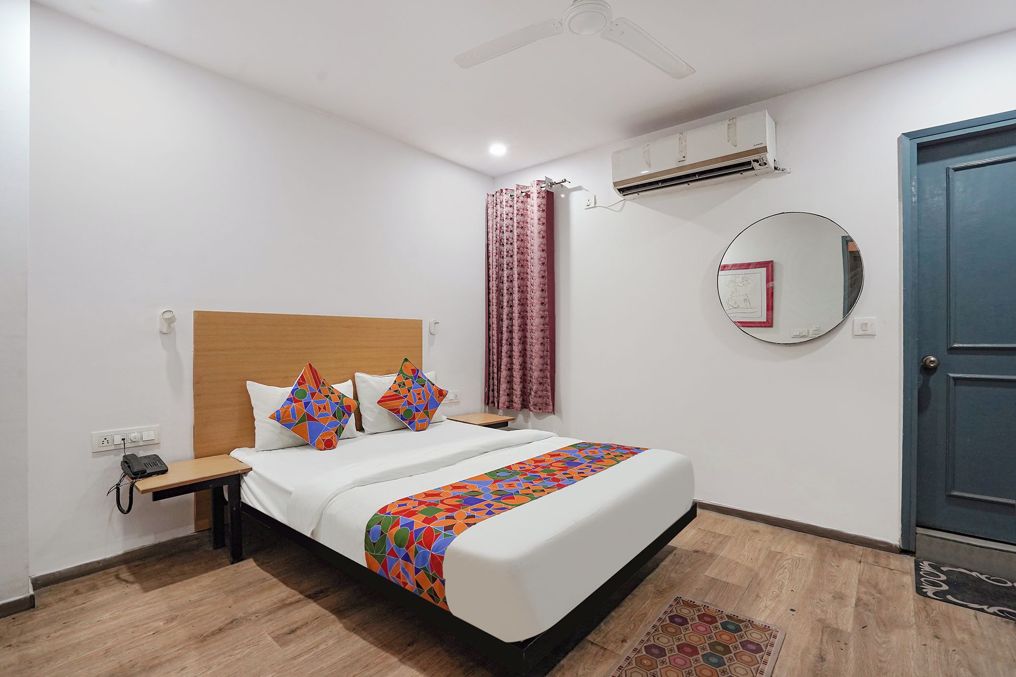 A hotel room with a bed, bedside table, phone, air conditioning unit, and a door.