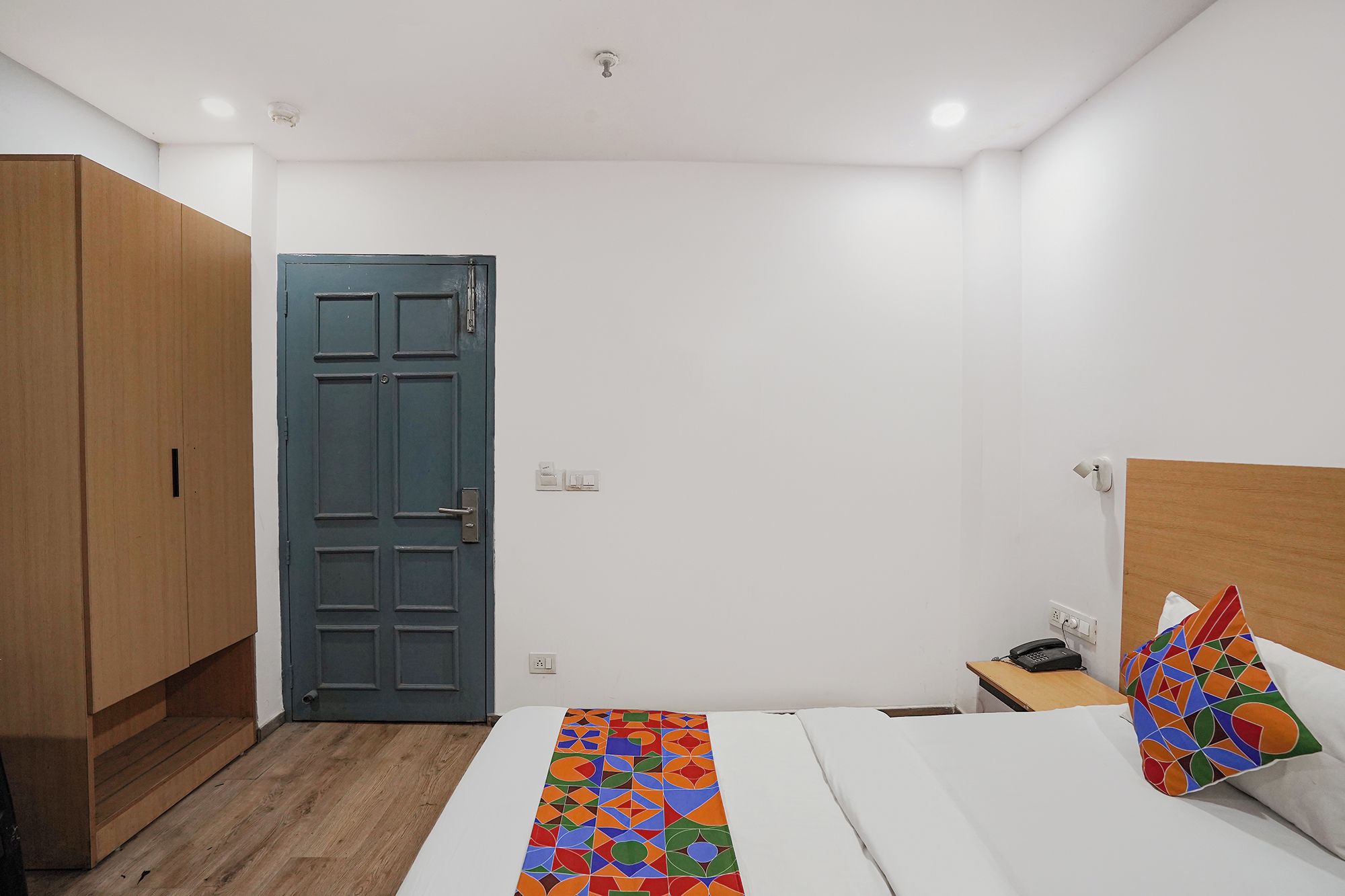 A hotel room with a bed, a wooden wardrobe, a blue door, a bedside table with a phone, and a colorful pillow.