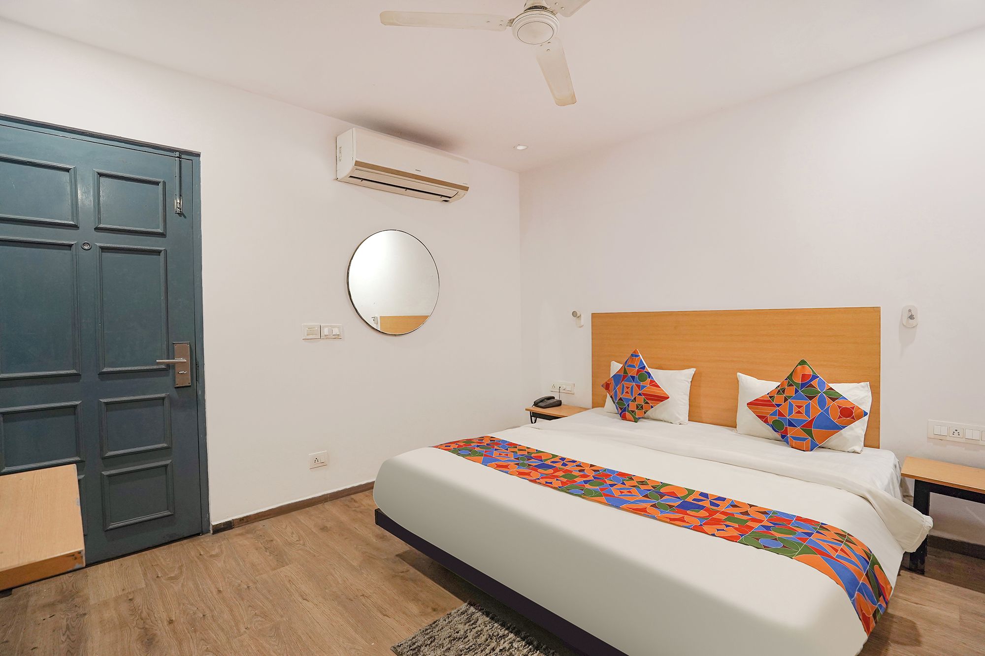 A hotel room with a bed, a nightstand, a bedside table, a ceiling fan, an air conditioning unit, a door, and a mirror.