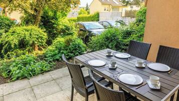Eine Terrassengruppe draußen mit Tisch für vier Personen, umgeben von Pflanzen und einem geparkten Auto.