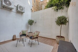 Ein kleiner Patio-Bereich mit Korbmöbeln, zwei Kübelbäumen und Klimaanlagen an der Wand.