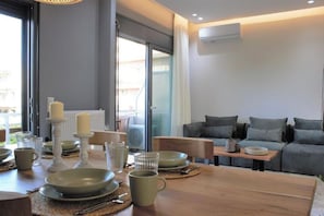 A modern living room with a sofa, a dining table set with plates, cups, and cutlery, and a large window providing natural light. - Koukounari Apartment in Downtown Chania (Chania)