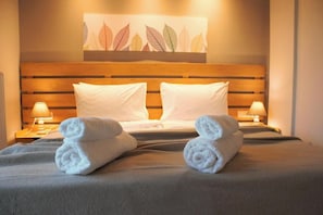 Ein Hotelzimmer mit einem hölzernen Kopfteil, zwei weißen Kissen, einem Bett mit einer grauen Tagesdecke, zwei zusammengerollten Handtüchern auf dem Bett und einem Wandbild über dem Bett.