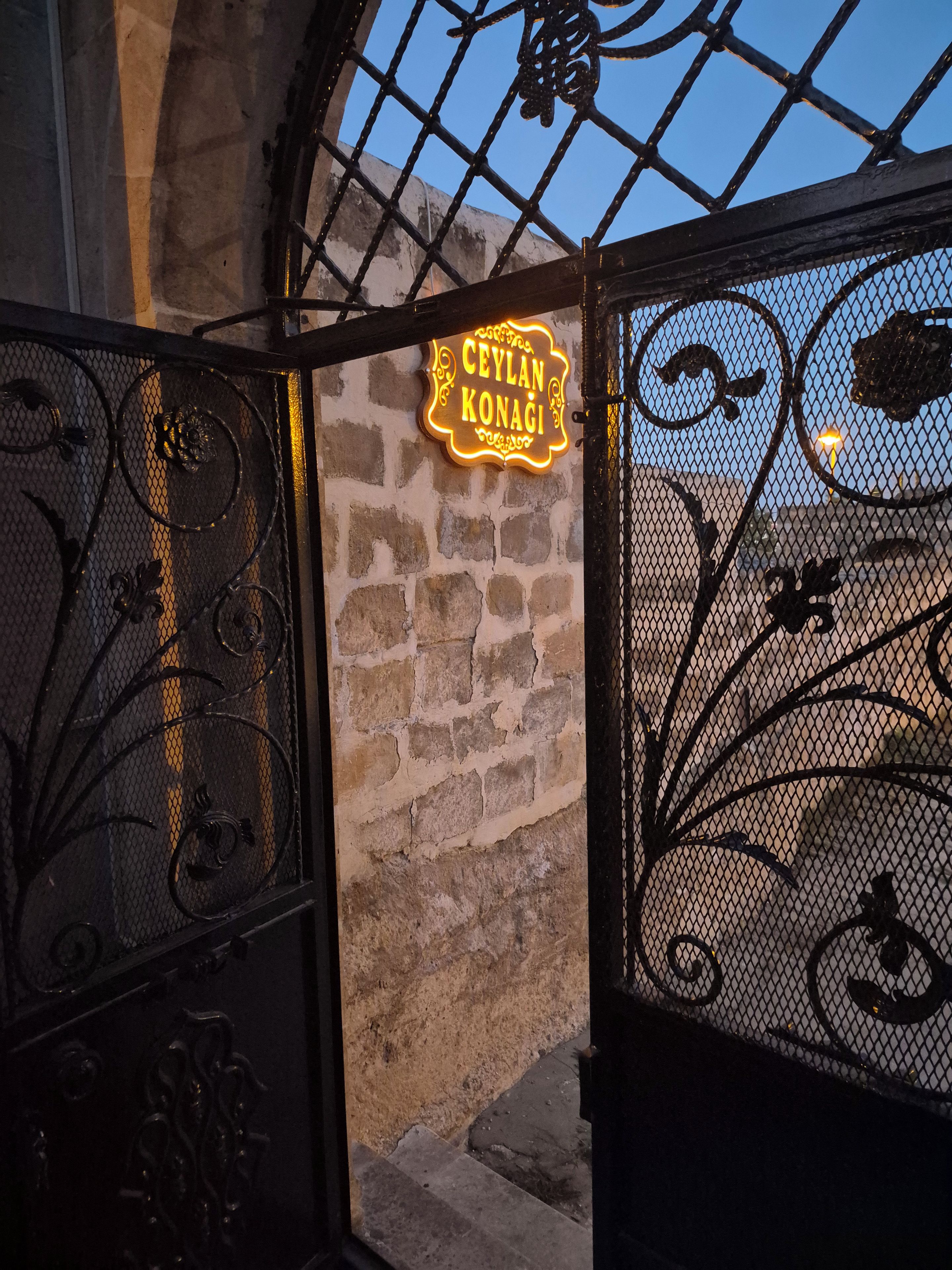 Ein schmiedeisernes Tor mit einem leuchtenden Neonzeichen, auf dem 'CEYLAN KONAGI' steht, vor einer Steinmauer.