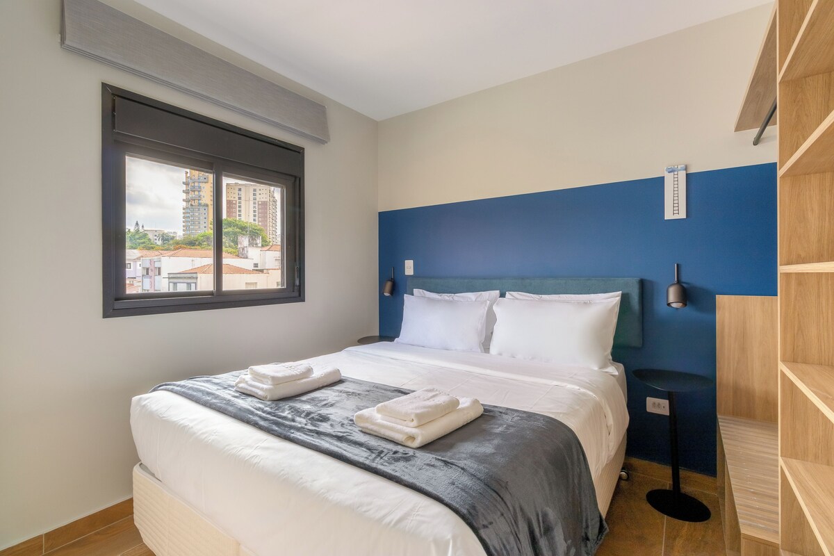 A hotel room with a bed, a window, a blue accent wall, and a view of buildings outside.
