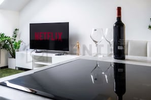 A modern living room with a flat-screen TV displaying 'NETFLIX' in red letters, a bottle of wine, and two wine glasses on a reflective surface. - Deluxe Riverside Duplex in the Heart of the City (Prague)