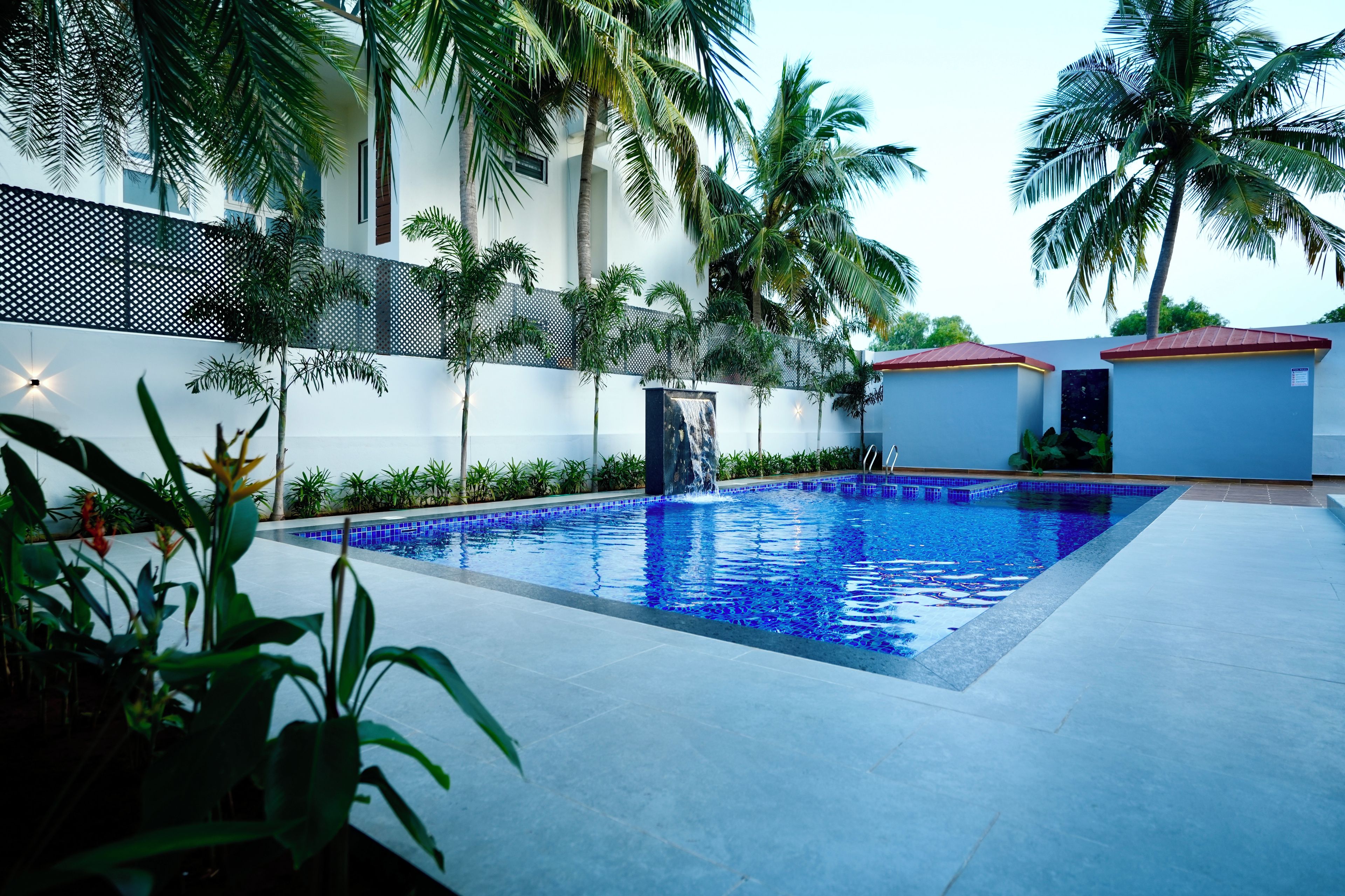 Una piscina con borde de azulejos, rodeada de palmeras y un edificio blanco con techo rojo.