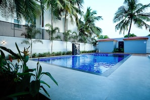 A swimming pool with a tiled edge, surrounded by palm trees and a white building with a red roof. - LA COAST (4 bedrooms) (Tirupporur)