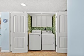 Une buanderie équipée de deux machines à laver et d’un sèche-linge, avec des placards blancs au-dessus et un mur décoré d’un motif de feuilles vertes.