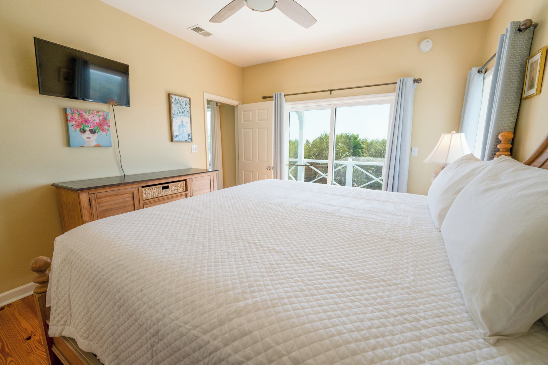 A bedroom with a bed, a flat-screen TV, a wooden dresser, and a balcony with curtains.