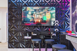 A room with a large screen displaying a video game, a gaming console, and two bar stools. - PROMO 5 BR Villa | Games | Pool | Near Disney - by Blue Sky Villas (Davenport)