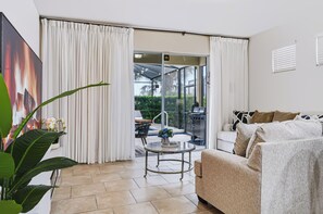 A modern living room with a glass door leading to an outdoor patio, a sofa, a small table, and a potted plant. - PROMO 5 BR Villa | Games | Pool | Near Disney - by Blue Sky Villas (Davenport)