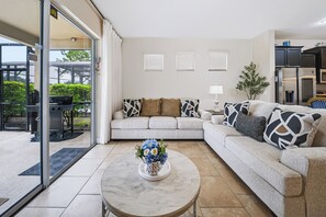Ein modernes Wohnzimmer mit einer Sofagarnitur, einem Couchtisch mit floraler Dekoration und einer Schiebetür zu einem Außenbereich.