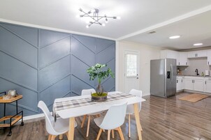Una cocina moderna con armarios blancos, un refrigerador de acero inoxidable y un comedor con mantel a cuadros y sillas.