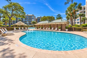 Una piscina con sillones de descanso y una zona de estar cubierta, rodeada de palmeras y un edificio de varios pisos.