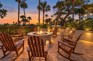 A patio area with a fire pit, outdoor seating, and a view of palm trees and a building in the background. - 4309 Windsor Court North | Oceanfront 4BR Villa + Sweeping Ocean Views (Hilton Head Island)