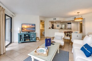 A modern living room with a television, a dining area with a table and chairs, and a kitchen in the background. - 3229 Villamare | Beach Access + Incredible Resort Pools (Hilton Head Island)