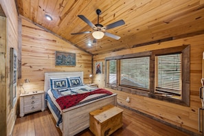 A wooden cabin bedroom with a bed, nightstands, a ceiling fan, and a window with blinds. - The Art Loft. A piece of history nestled a mere quarter-mile from the Smoky Moun (Sevierville)