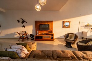 Ein Wohnzimmer mit einer Couch, einer Holztrennwand mit Fernseher, einem Korbstuhl und einem kleinen Tisch mit Ventilator.