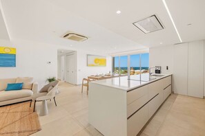 A modern kitchen with a central island, a dining area with a view, and a living space with a sofa and chairs. - A360.W31 - Tropical Vista 3BR Suite (Noord)